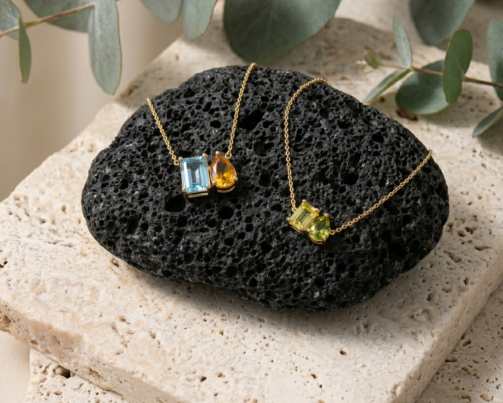 Two gold necklaces with gemstones on a black stone surface with green leaves in the background